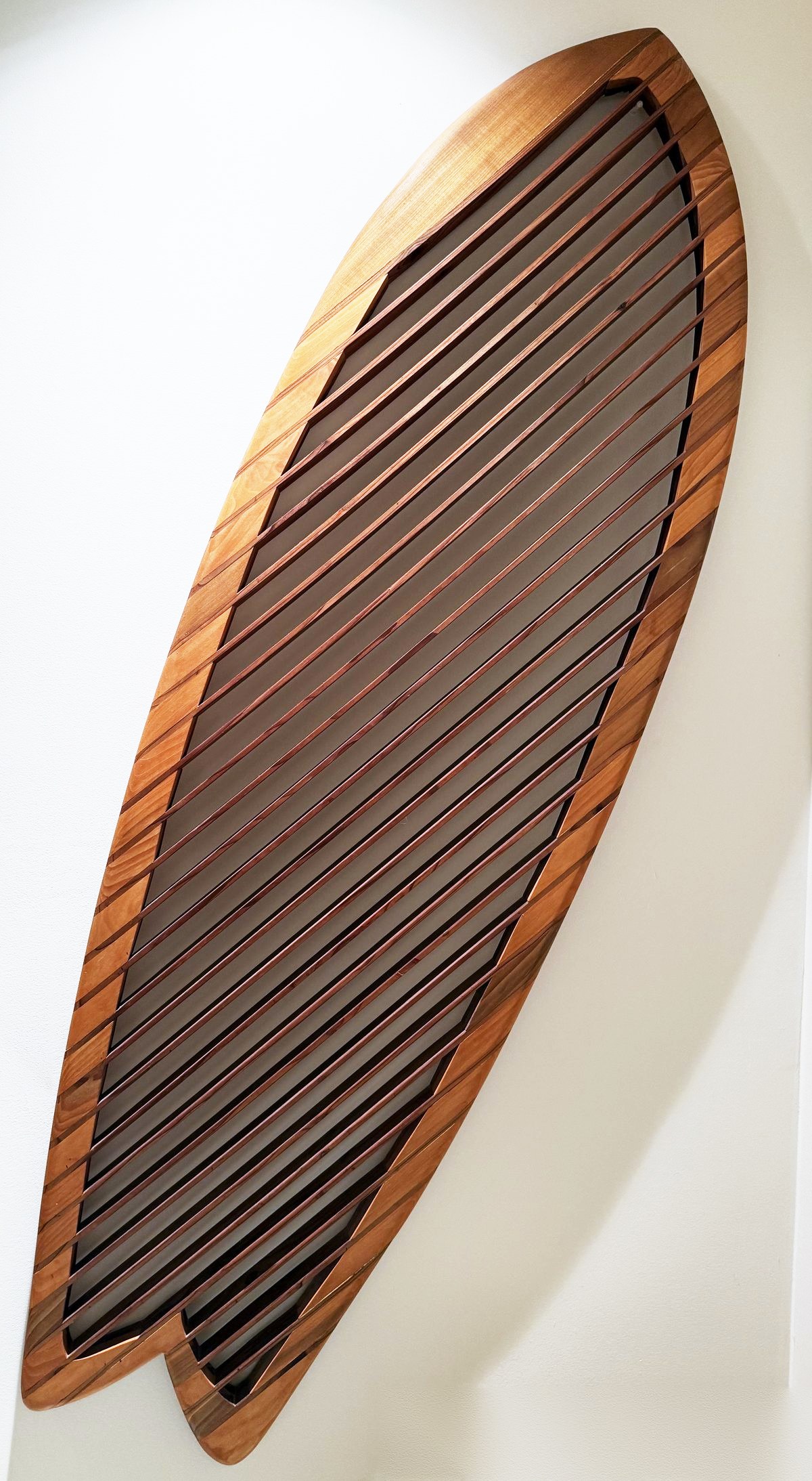 Alaia: a twin-fin surfboard form with laminated cedar rails and diagonal redwood slats, mounted on a white wall.